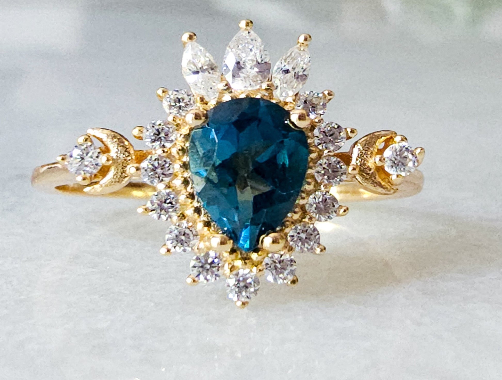 A pear-shaped London Blue Topaz gemstone with cubic zirconia accents around the stone on a gold-plated ring sitting on a marble table.